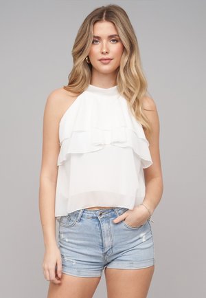 Junge Frau mit langen blonden Haaren, die ein ärmelloses weißes Rüschentop und hellblaue, ausgefranste Denimshorts trägt, steht mit einer Hand in der Tasche.