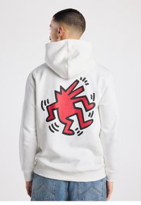 Witte hoodie met een oversized rood-zwart grafisch ontwerp op de achterkant, met een gestileerde figuur en dynamische lijnen. Zachte stof, standaard pasvorm.