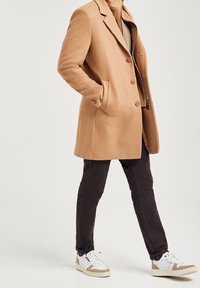 Beige ullrock med skräddarsydd design, två knappar och sidfickor. Matchad med mörka byxor och vita sneakers med beiga detaljer.