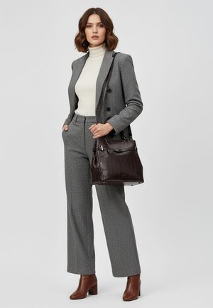 Femme en tailleur gris, col roulé blanc, bottines marron, tenant un sac à main en cuir marron foncé sur l'épaule.