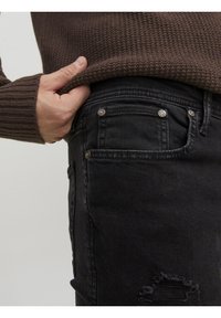 Ręka podciągająca brązowy, teksturowany sweter nad kieszenią czarnych, podartych dżinsów z nitami na jasnym tle.