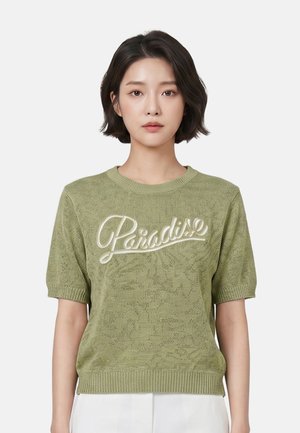 Vrouw met kort donker haar draagt een lichtgroene gebreide top met korte mouwen met witte tekst "Paradise,", gecombineerd met een witte broek, neutrale achtergrond.