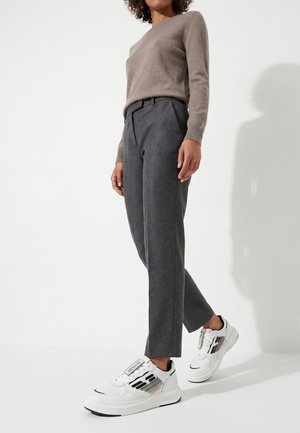 Personne portant un pantalon gris taille haute, un pull taupe à manches longues et des baskets blanches avec des détails noirs et beiges sur un fond blanc.