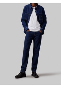 Muž oblečený v tmavě modré džínové bundě a kalhotách s bílým tričkem a černými koženými šněrovacími botami, ruce v kapsách, stojící na šedém pozadí.