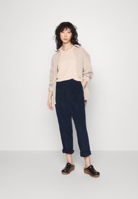 Camicia beige abbottonata sopra un top beige chiaro, abbinata a pantaloni di velluto a coste blu navy e zoccoli neri con suola di legno, con i pantaloni arrotolati.