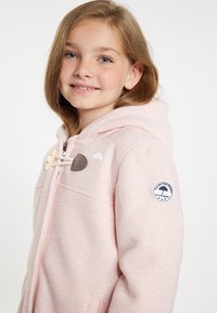 Felpa con zip di colore rosa chiaro, realizzata in tessuto texturizzato, con chiusura a toggle in legno e morbidi risvolti grigi e bianchi; presenta una toppa con logo sulla manica.