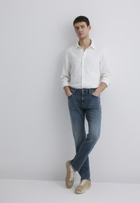 White button-up shirt, blue denim jeans, and beige sneakers. The shirt has rolled sleeves and a classic collar; jeans are slightly fitted.