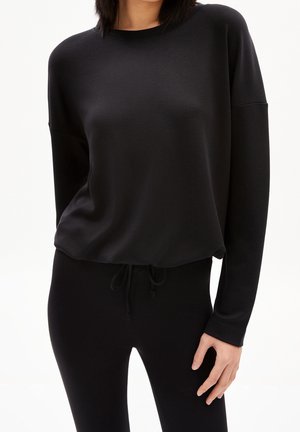 Vrouw draagt een effen zwarte top met lange mouwen en een ronde hals, gecombineerd met zwarte broek met trekkoord, staand tegen een witte achtergrond.