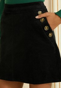 Tuvo skatījums uz cilvēku, kurš valkā melnu kordūrskreklu ar četriem lieliem pogām un roku, kas atpūšas sānu kabatā.