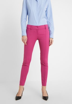 Vrouw draagt strakke felroze broek, lichtblauw shirt met lange mouwen en zwarte pumps, staand tegen een effen achtergrond.