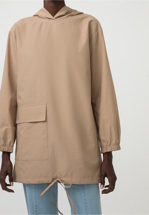 Person iført en beige hættejakke med en stor forlomme og snøre i kanten, kombineret med lyseblå jeans mod en ensfarvet baggrund.
