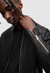 Schwarze Lederjacke mit silbernen Reißverschlüssen, steigendem Kragen und tailliertem Design. Das Model berührt den Kragen und zeigt die Textur und Details.