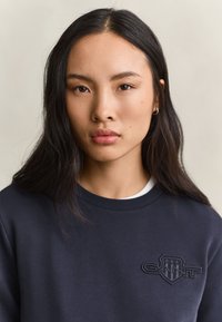 Jonge vrouw met lang zwart haar draagt een donker marineblauwe sweatshirt met een schildlogo en het woord "GANT" op de borst, tegen een effen achtergrond.
