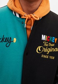 Veste universitaire colorée en laine turquoise et noire, col rayé, détails brodés multicolores, avec un sweat à capuche orange en dessous.