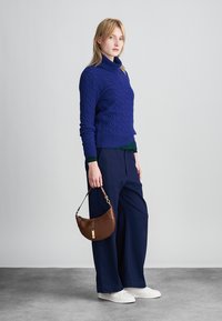 Μπλε πουλόβερ με πλέξη καλωδίων και ψηλό γιακά, συνδυασμένο με ναυτικά παντελόνια σε χαλαρή γραμμή. Κρατάει μια καφέ δερμάτινη τσάντα ώμου, στέκεται σε μία υφασμένη επιφάνεια.