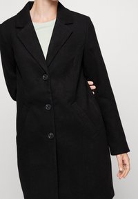 Cappotto di lana nera con colletto a revers, design a un solo petto e quattro bottoni; presenta tasche laterali e una finitura liscia e testurizzata.