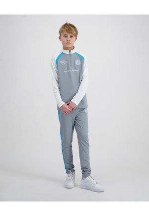 Jongen staat met gevouwen handen, draagt een grijs-wit trainingspak met blauwe strepen en witte sneakers tegen een effen lichte achtergrond.