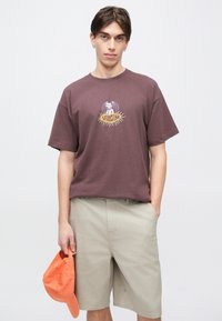 Νέος άνδρας φορώντας σκούρο μωβ μπλουζάκι με σχέδιο και μπεζ σορτς, κρατώντας πορτοκαλί καπέλο του μπέιζμπολ στο αριστερό του χέρι.