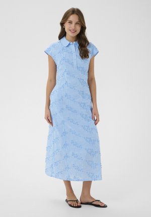 Donna in piedi, indossa un vestito midi azzurro chiaro senza maniche con motivo floreale, colletto e sandali neri piatti, sorridente con i capelli sciolti.