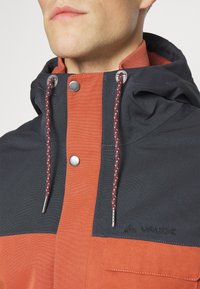 Giacca con cappuccio nero e arancione, caratterizzata da un tessuto testurizzato, chiusure a pressione in metallo e cordini regolabili con dettagli decorativi.