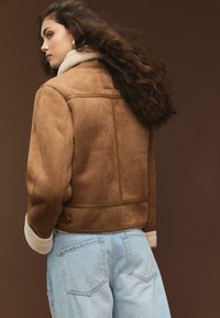 Brun mockaskinnjacka med fårskinnskrage, rektangulär form och sidofickor, i kombination med ljusblå denimjeans med bakfickor.