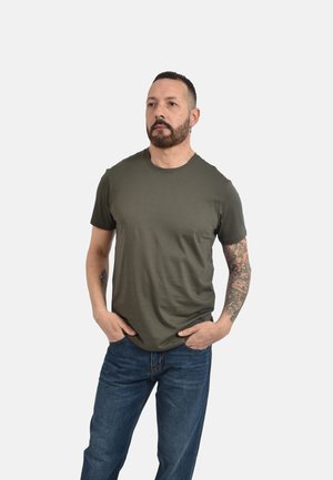 Uomo con barba e tatuaggi che indossa una maglietta verde e jeans blu, in piedi con le mani nelle tasche davanti a uno sfondo semplice.