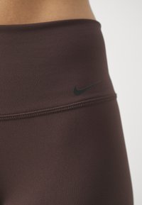 Hnědé sportovní legíny s vysokým pasem, hladkou texturou a černým logem Nike na pase. Obsahují bezpečné šití.