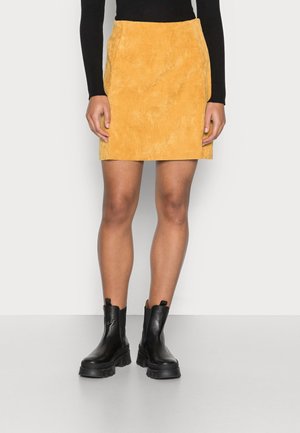 Femme portant une mini-jupe en velours côtelé jaune moutarde, un haut noir à manches longues et des bottines en cuir noir épaisses, debout sur un sol gris.