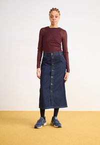 Μίντι τζιν φούστα με κουμπιά μπροστά, συνδυασμένη με ένα μπορντό μπλουζάκι μακρυμάνικο και μπλε αθλητικά παπούτσια. Το φόντο έχει ουδέτερο τόνο.