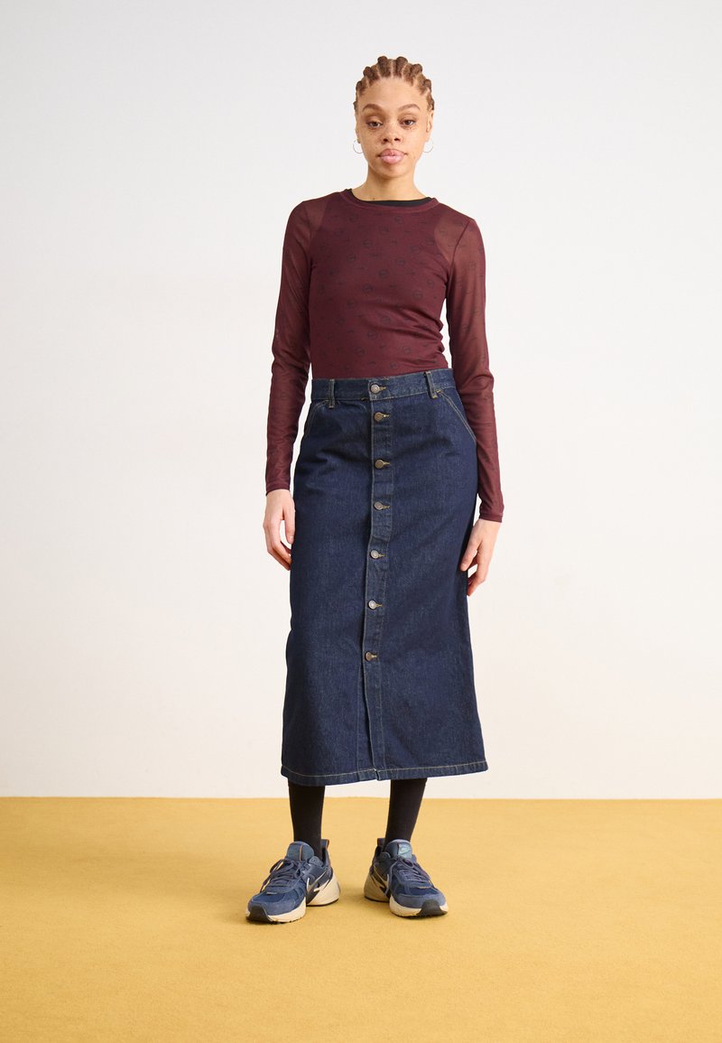 Denim midi rok met knopen aan de voorkant, gecombineerd met een bordeaux rood langemouwen top en blauwe sneakers. De achtergrond is in een neutrale tint.
