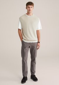 Tânăr în picioare, purtând un pulover bej fără mâneci peste un tricou alb, pantaloni gri, pantofi negri și un ceas de mână.