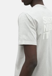 Camiseta de algodón gris con cuello redondo, que presenta un atrevido texto blanco que dice "SOSTENIBILIDAD". Etiqueta "ECOALF" mostrada en la manga izquierda.