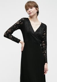 Svart kjole med lange blondeermer, V-hals og flytende skjørt. Blonden har et blomstermønster, som står i kontrast til det solide stoffet.