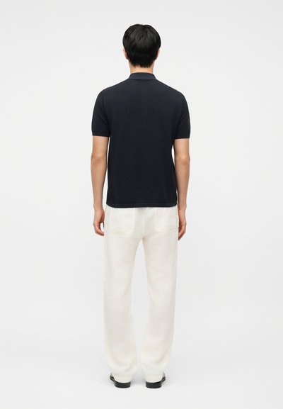 Homme de dos portant un polo noir à manches courtes, un pantalon blanc et des chaussures noires sur un fond blanc uni.