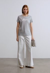 Γκρι κοντομάνικη μπλούζα με το κείμενο «Bae», συνδυασμένη με λευκό παντελόνι φαρδιάς γραμμής και μπεζ παπούτσια. Το μοντέλο κρατά μια ανοιχτό γκρι τσάντα.