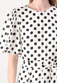 Vestido branco com bolinhas pretas, com mangas bufantes, gola redonda e cintura ajustada com um detalhe atado. Tecido liso e leve.