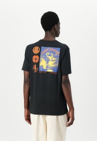 Tricou negru din bumbac cu un print grafic pe spate în galben și portocaliu, cu detalii ale trupei și logo-ului. Design cu mâneci scurte și gât rotund.