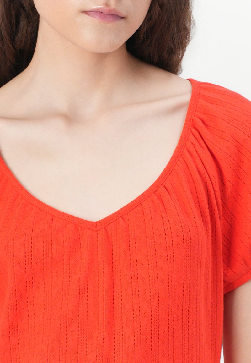 Rote Bluse mit V-Ausschnitt, kurzen Puffärmeln, strukturiertem Rippstoff und subtilen vertikalen Linien. Nahaufnahme des Oberteils.