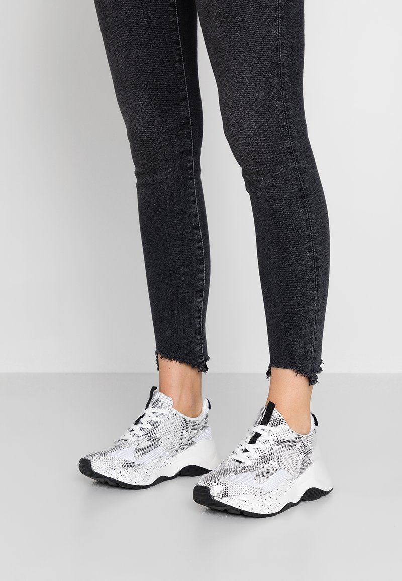 Personne portant des baskets épaisses blanches et noires avec un motif serpent, associées à un jean skinny noir à ourlet effiloché sur un sol uni.