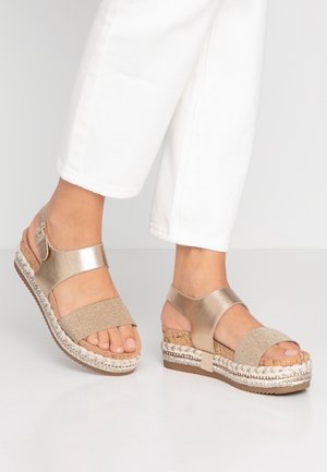 Feet wearing gold metallic wedge sandals with cork and braided rope soles, paired with white cropped pants on a white floor.
