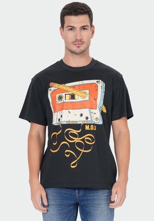 Man draagt zwart T-shirt met een grote afbeelding van een oranje-wit cassettebandje met uitgerolde tape en de tekst "M.O.D".
