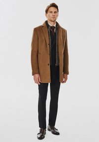 Brązowy wełniany płaszcz o usztywnionej konstrukcji, nałożony na czarną kamizelkę i zapinaną koszulę, połączony z czarnymi spodniami i ciemnymi butami.