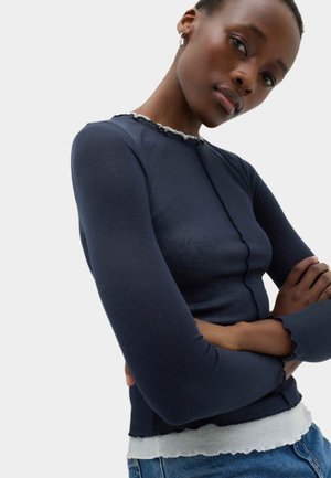 Ung person med mørk hud iført en tætsiddende navyblå langærmet top og lyseblå jeans, armene krydsede, kigger lidt nedad.