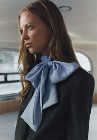 Pajarita de seda azul claro atada al cuello, llevada sobre un blazer de cuadros oscuro. Cabello largo y liso cae sobre el hombro. Iluminación suave.