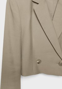 Veste beige à double boutonnage avec un design structuré, des revers crantés et deux grands boutons transparents, présentant une texture lisse et tissée.