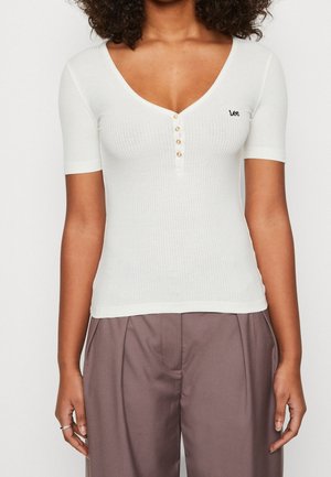 Vrouw draagt een wit geribd kort mouwtopje met vier knopen en een klein "Lee"-logo op de borst, gecombineerd met losse bruine broek.