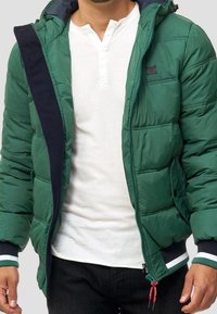 Giubbotto a forma di piumino verde con cappuccio, presenta una zipper e dettagli blu navy, polsini elasticizzati e orlo con cordino. Indossato sopra una maglietta bianca a maniche lunghe.