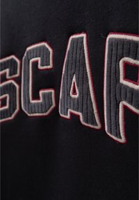 Tessuto nero con grandi lettere grigie ricamate in rilievo e contornate di rosso. Le lettere sono in uno stile di font sportivo con cuciture visibili.