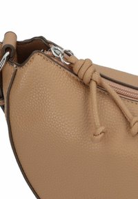 Bolso de cuero color marrón claro con textura granulada, que presenta una forma redondeada, un cierre de cremallera y un acento con cordón anudado. Hardware en plateado incluido.