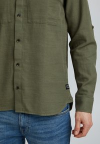 Camicia verde oliva con bottoni, due tasche sul petto, maniche lunghe e spacchi ai lati. Il tessuto appare leggero con una texture sottile.